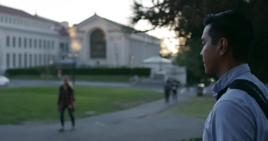 A young man with a laptop bag over his shoulder is walking along a path through a university setting