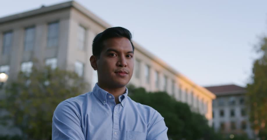 A young man in a blue button up shirt stands in front of a large building
