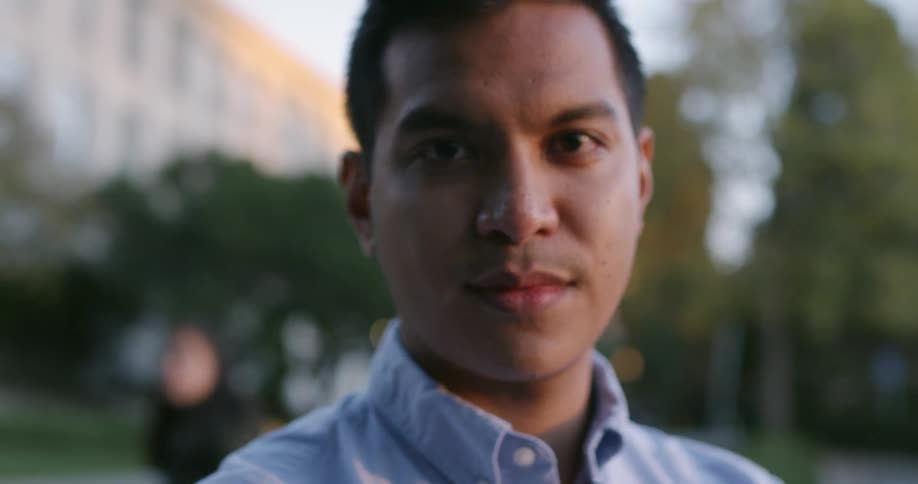 A young male standing next to a street looks off into the distance before turning his gave directly at the camera