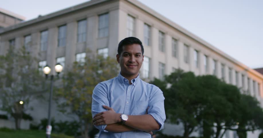 A man in a dress shirt smiles at a camera while standing in front of a building