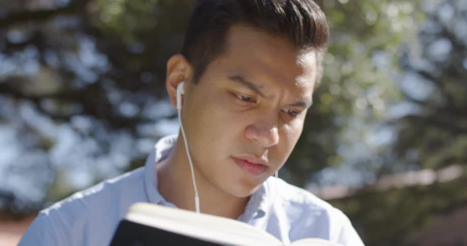 A man reads a book outside while wearing earbuds on a sunny day