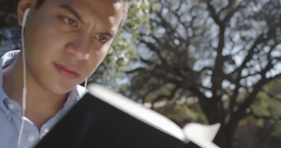 A hispanic man reads a book while wearing ear buds
