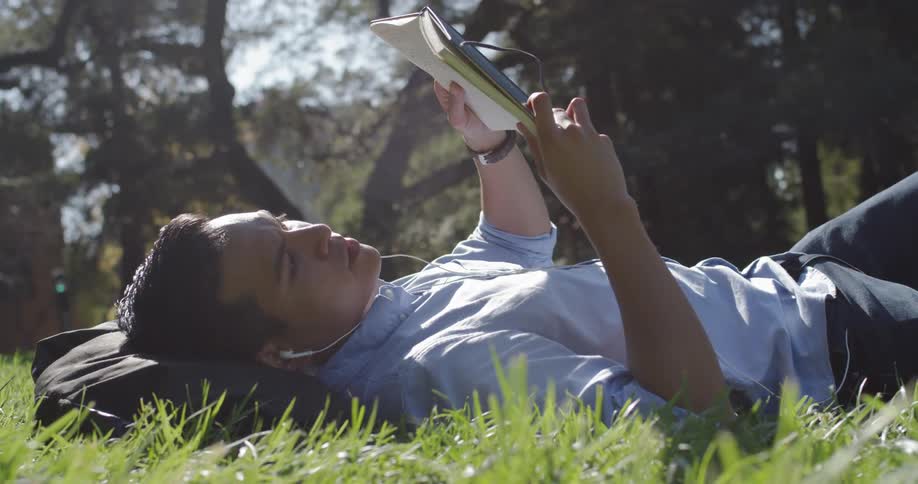 A man lies down on grass while reading a book outside on a sunny day