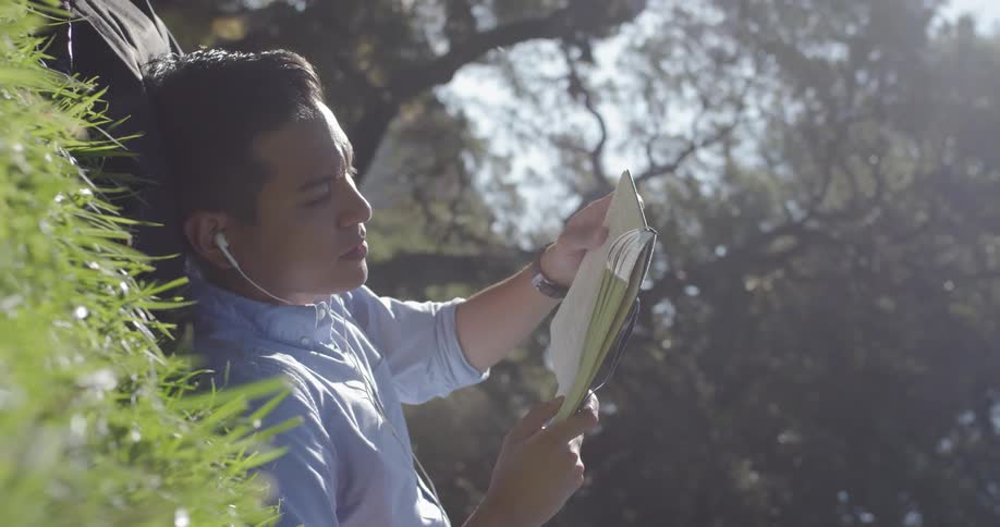 A man is laying on the grass reading a book with white earbuds in