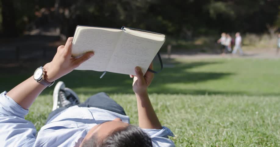 Man lies down on the lawn and flips pages of a notebook in the public park on a sunny day