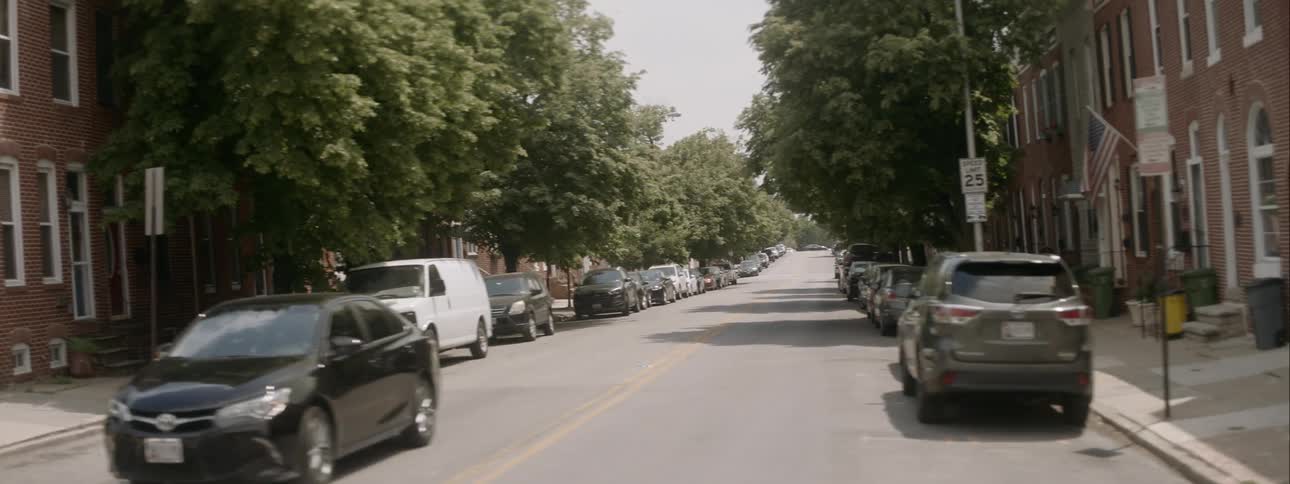 Passing by a car waiting at a stop sign on a side street with trees parked cars and brick face buildings