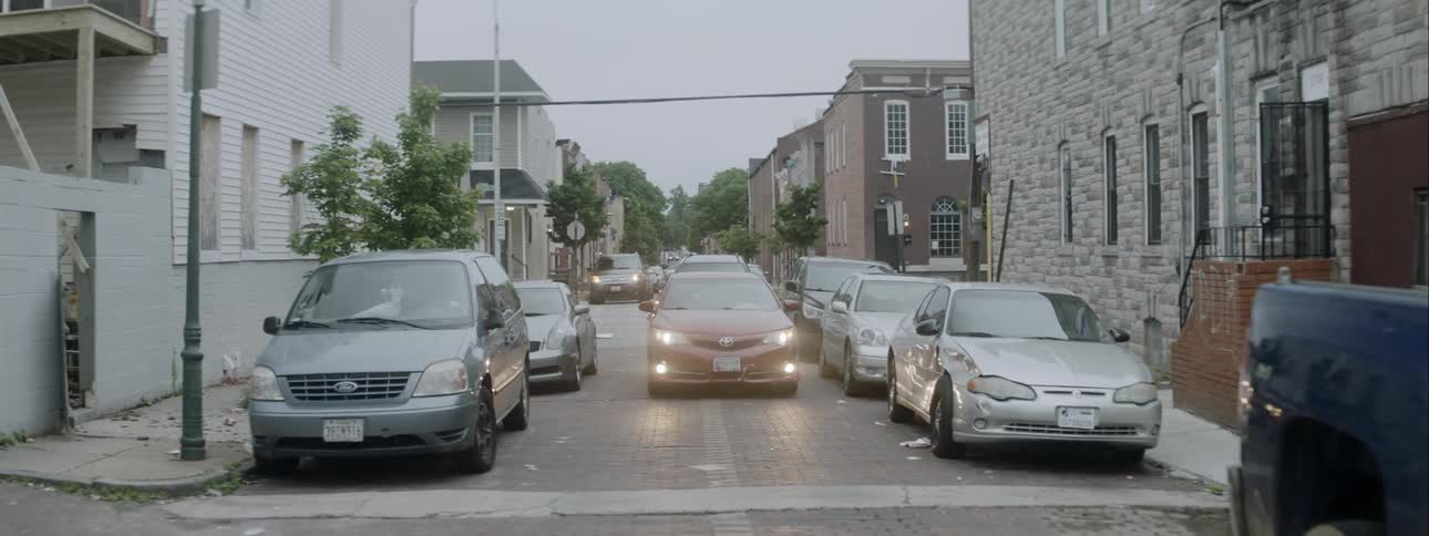Red car driving through neighborhood with many parked cars on street