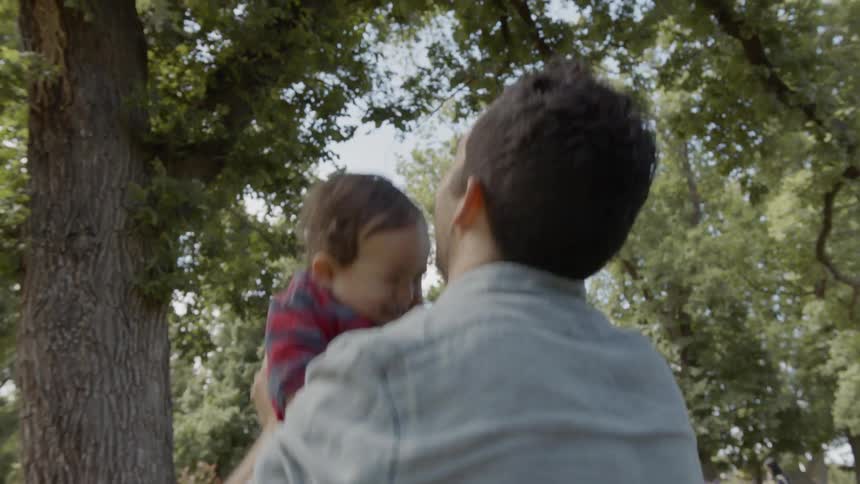 A man is tossing a baby in the air and catching him to make him laugh beneath a tall tree