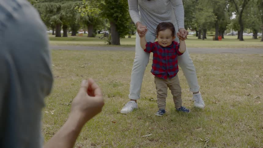 A mom and dad teach a toddler how to walk in a park