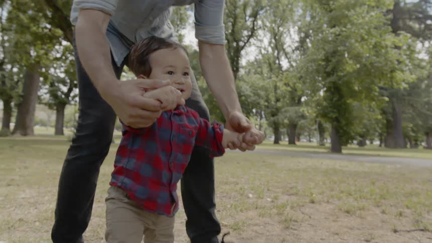 A man holding a babys hands to keep him balance as they walk in a park