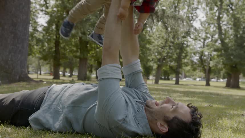 A man lays in the grass and raises a baby above him playing
