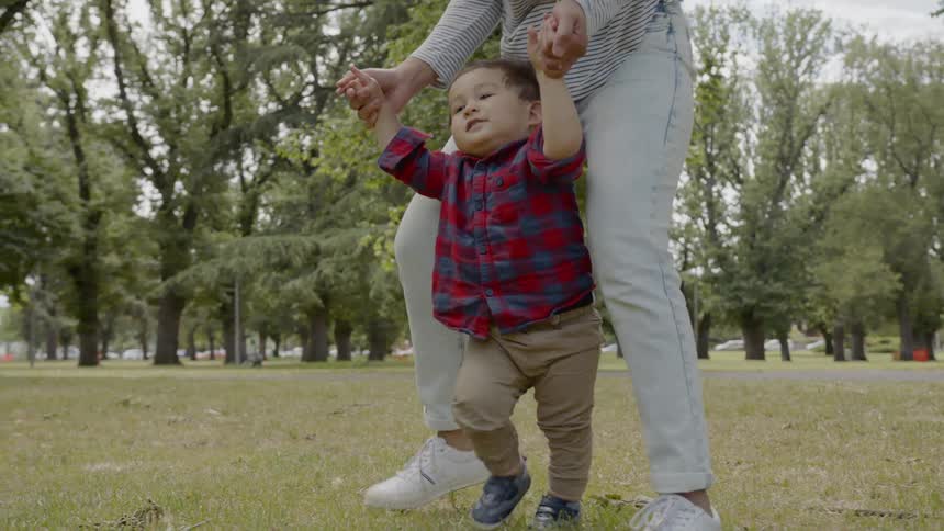 Woman helps toddler walk on lawn in the public park during daylight