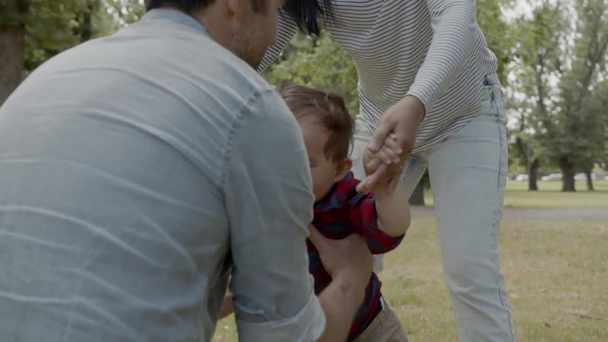 A little boy is walking towards his father while holding his mothers hands and the father picks him up