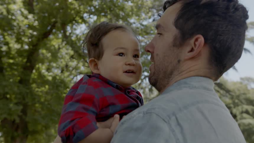A man is throwing a baby up in the air and catching him with tall trees in the background