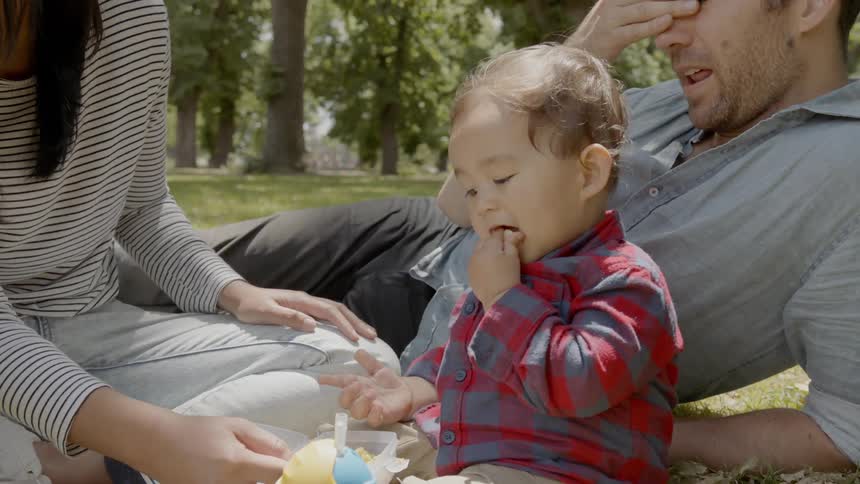 A young family sits on a grassy lawn and eats snacks