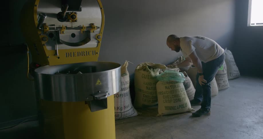 A man looks at bags of coffee next to a large coffee roaster in a factory