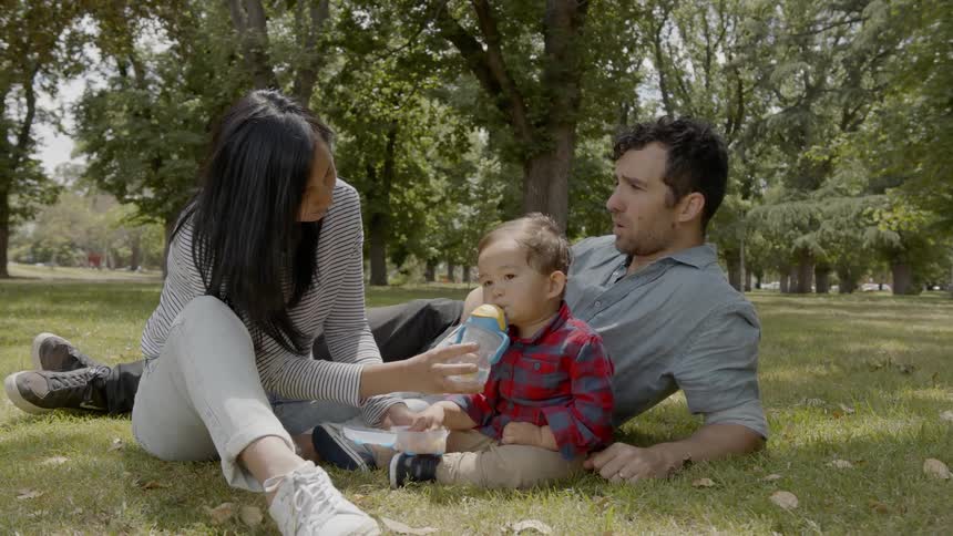 A family consisting of a man woman and child are sitting in the grass at a park while the female helps the baby drink from a straw
