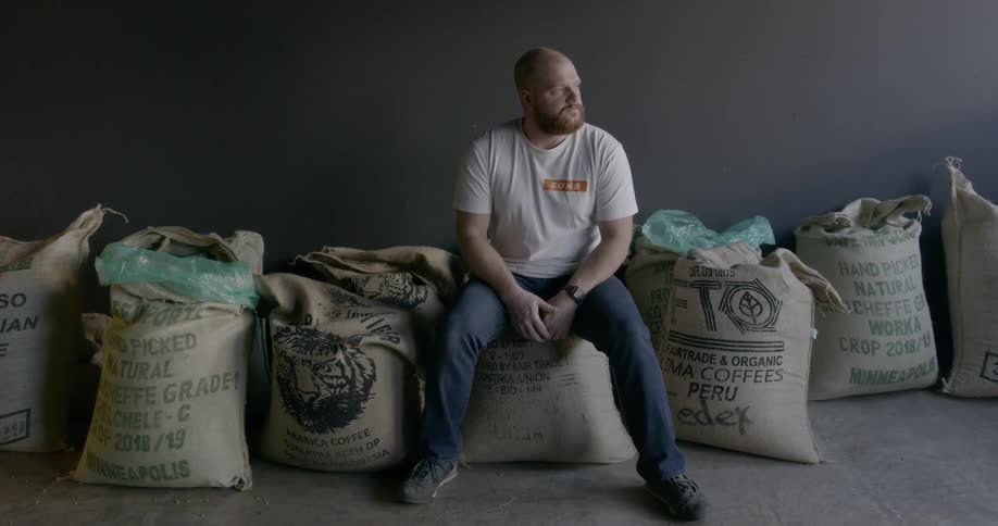 A man sits on a bag of coffee beans surrounded by other bags