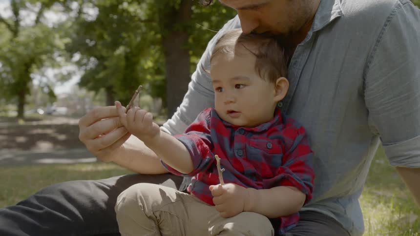 A young boy is playing with sticks while sitting on his fathers lap in the grass at a park