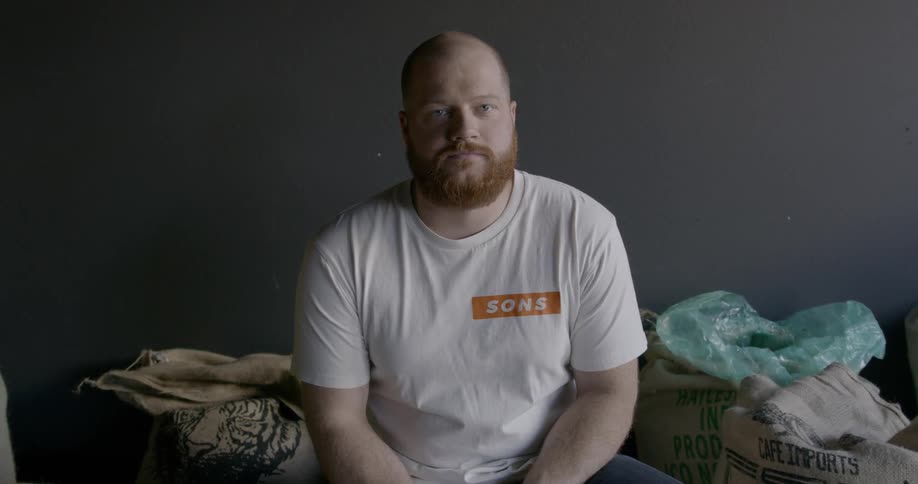 A bearded man is sitting on a bed and turns his head and smiles