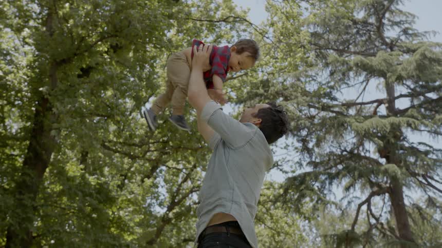 A man reaches down to pick up his son throws him into the air several times kisses him on the cheek and then hugs him to his shoulder