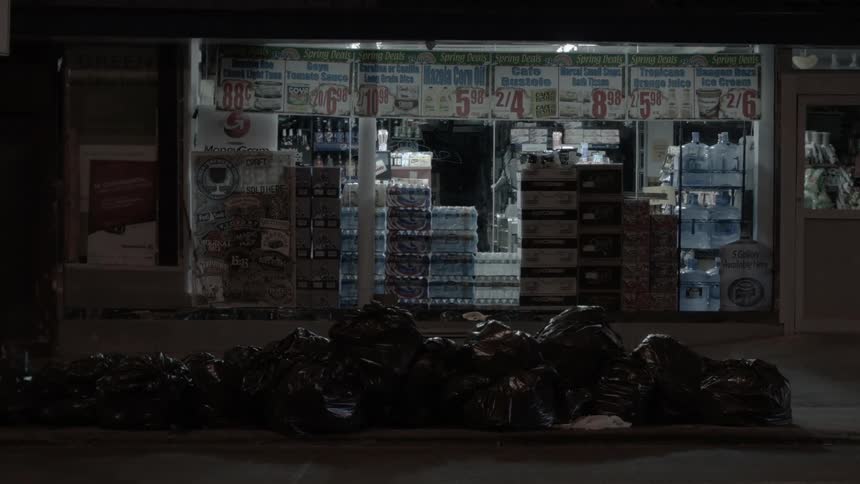 A pile of black garbage bags in front of a store with a white car driving in the foreground