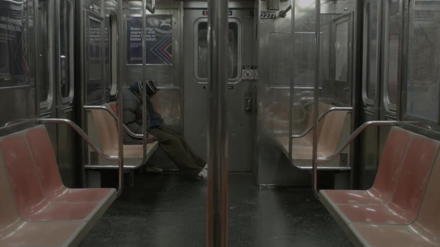 A person bundled in a winter coat and hat is sleeping in a seat in an otherwise empty subway car