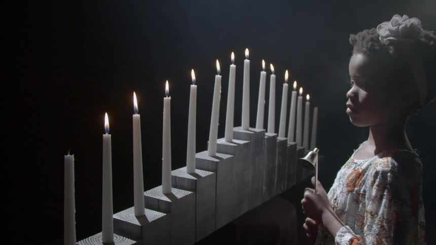 A young black girl holds a metal candle snuffer as she looks upon a row of candles aligned on pedestals in a darkened room