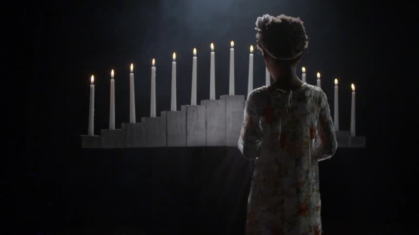 A female child approaching acandelabra of fifteen lit candles in a dark and misty room