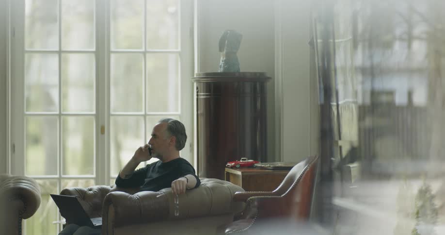A man sits on the couch holding his glasses and talking on the phone