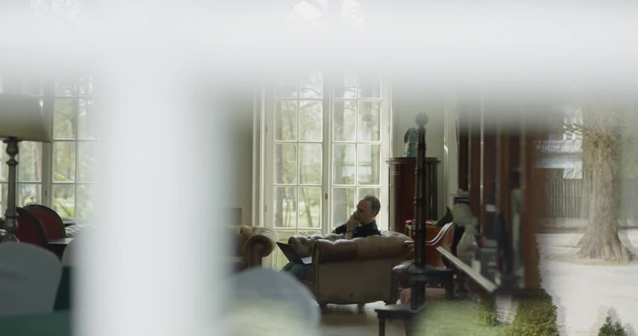A man sits in a chair with a laptop on his knee and talks on the phone