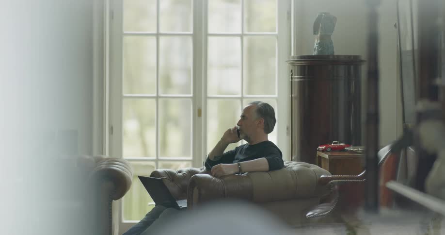 A man is sitting while he talks on the phone in his living room