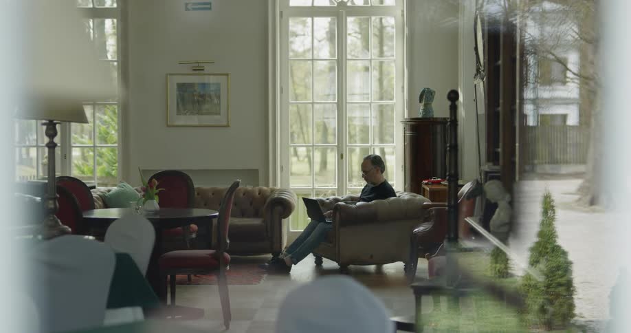 A man sits in a nicely furnished living room while working on his laptop in front of a large window