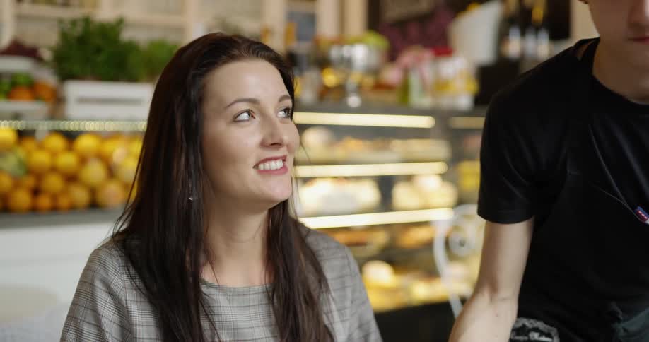 A server brings two large plated desserts to a woman sitting at a table talking to someone off screen