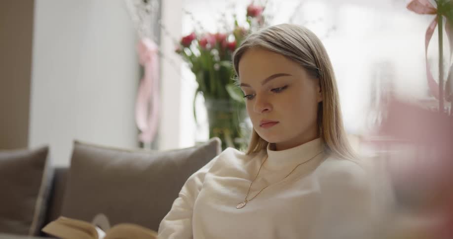 A blonde white woman sits on a couch and leafs through a book