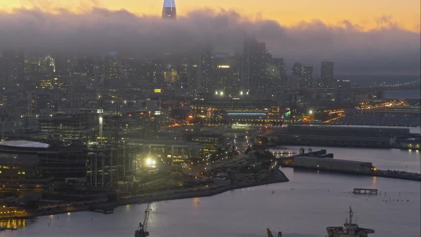 Buildings in a city are illuminated in the evening sky as clouds pass skyscrapers with one appearing above the clouds