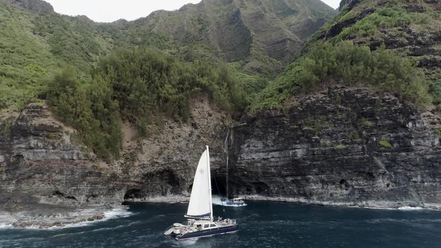 A boat drives through a tunnel with a waterfall and into a large lagoon past a sailboat