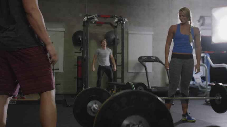 Muscular woman and man lift barbell in a gym with people working out in the background during daylight