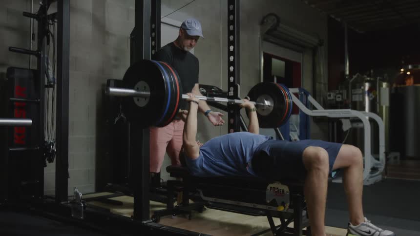 A man bench presses while another man stands above him and spots for him