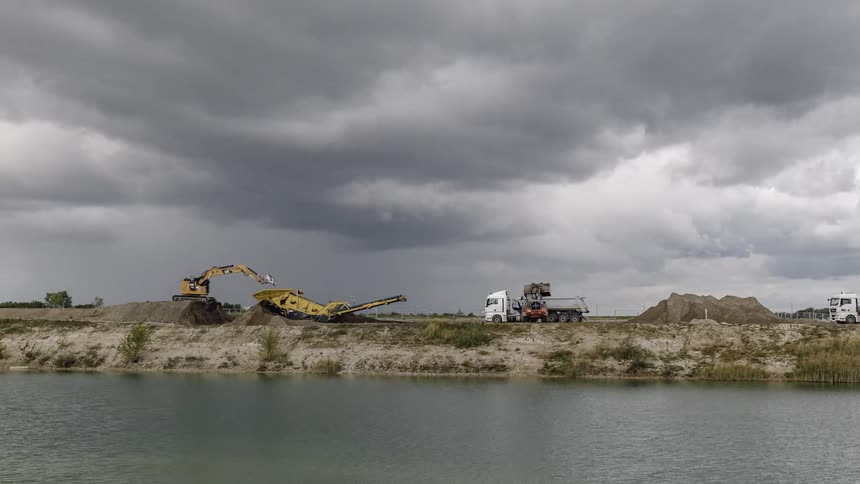 A loader fills dump trucks with soil while a backhoe digs on a cloudy day by the river