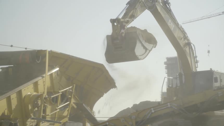 A large escavator digs dirt then puts it into a hopper which spits it onto a conveyor belt at a work site under a cloudy sky