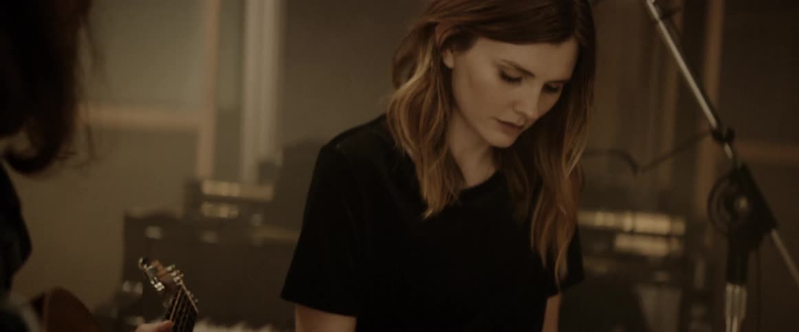 A man plays guitar while a woman writes lyrics in a recording studio