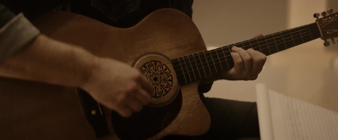 A young man plays guitar while a woman writes lyrics in a studio with closed blinds