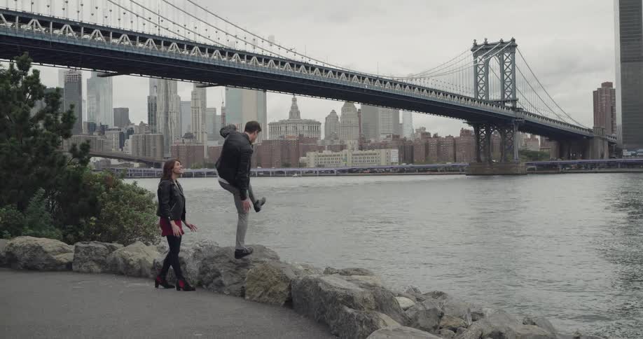 A man walking with a woman jumps up on a boulder and points out into the city harbor