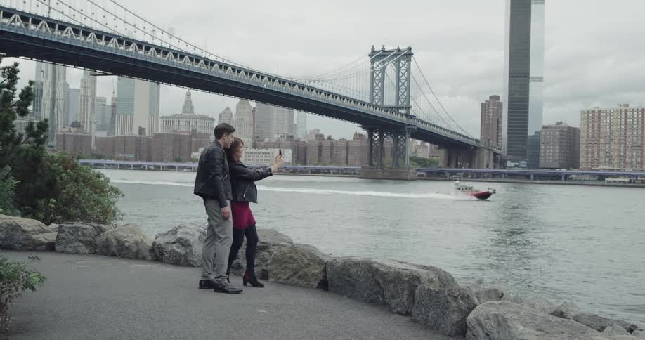 Woman and man take a selfie with smart phone on the promenade by the riverside across new york city and manhattan bridge during daylight and b