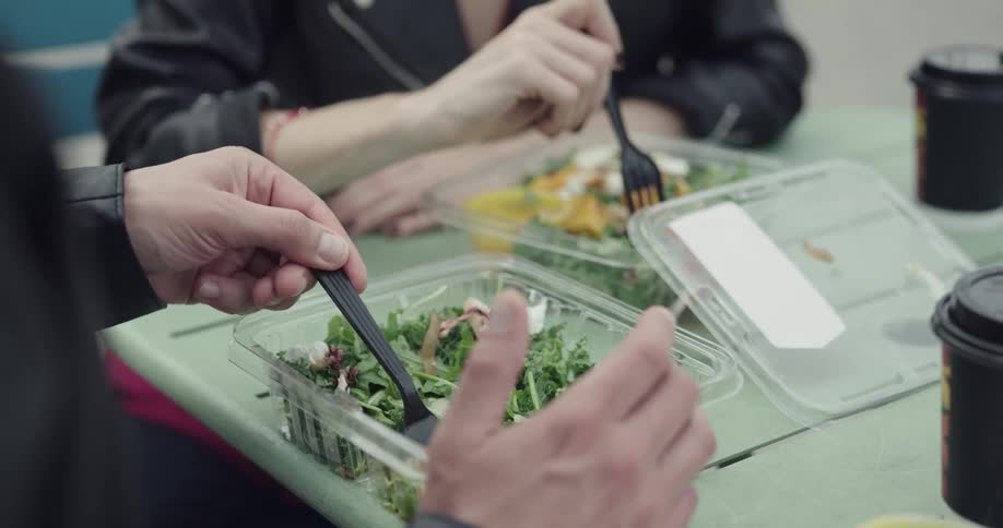 A man and woman are eating salads from plastic containers on a small outside table
