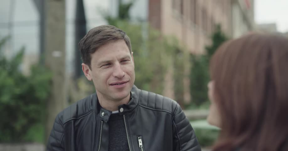 A man and woman smile while talking outside in a city