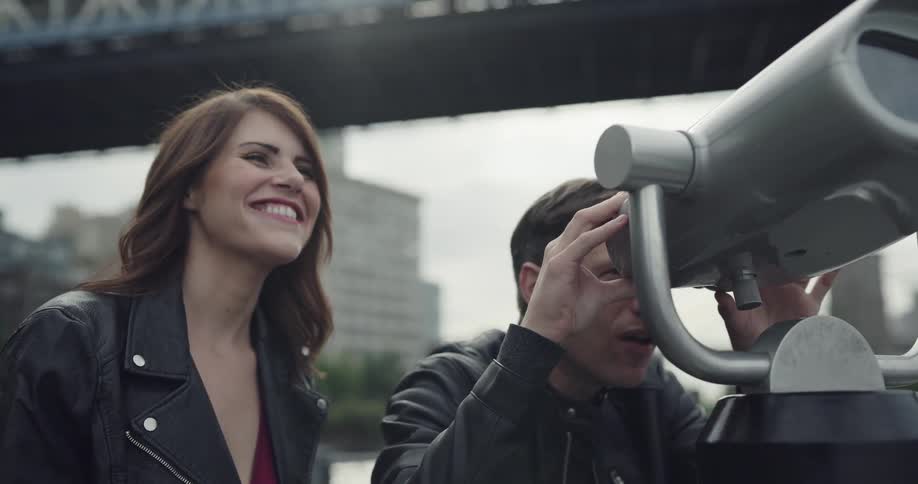 A man looks in a telescope as a woman stands beside him smiling and laughing