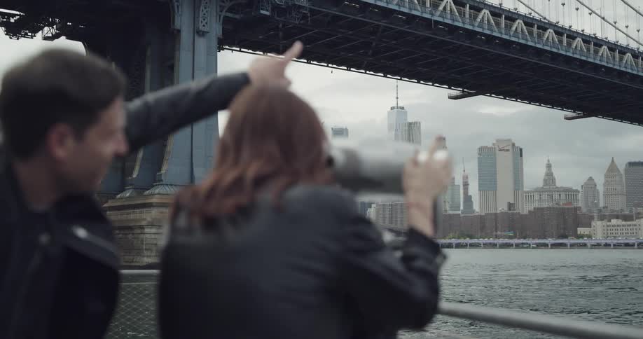 A man and woman look through public binoculars in a park across from new york city