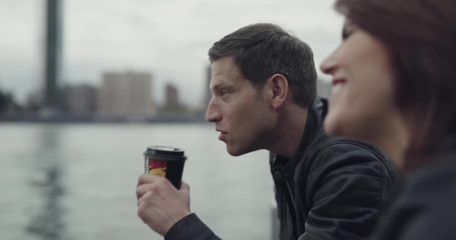 A man points while talking to a smiling woman next to a rippling river in a city